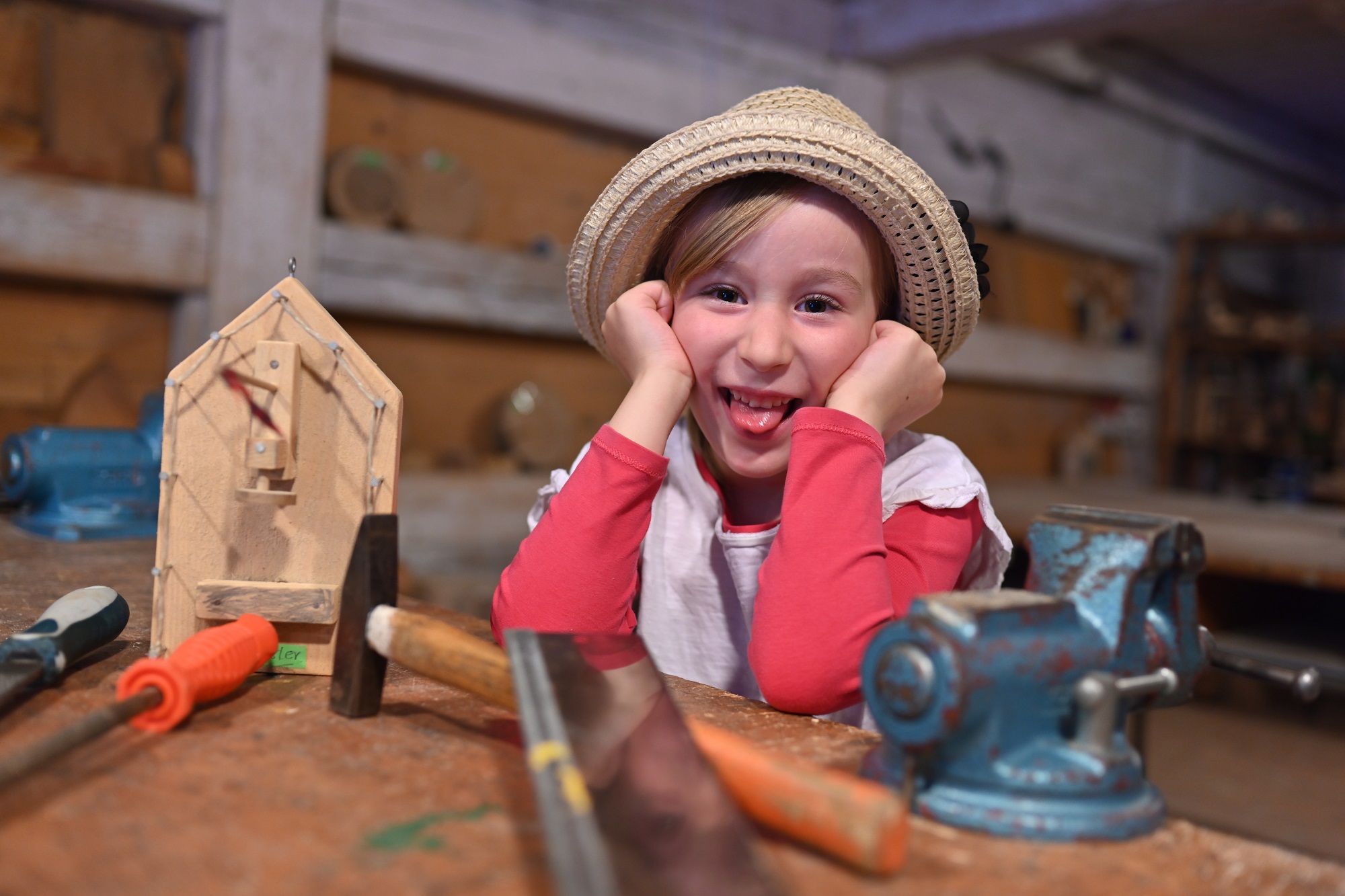 Kind sitzt in der Holzwerkstatt mit Werkzeug und Holzhaus